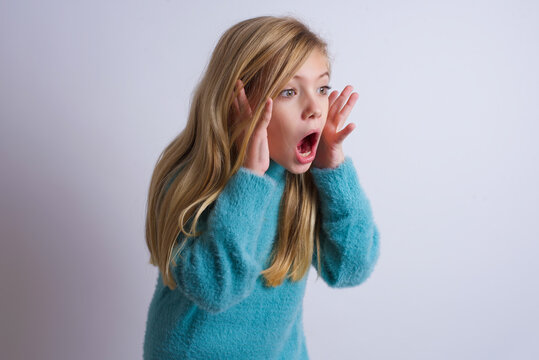 Cute Caucasian kid girl wearing blue knitted sweater against white wall with shocked facial expression, holding hands near face, screaming and looking sideways at something amazing.