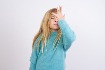 Frustrated Cute Caucasian kid girl wearing blue knitted sweater against white wall holding hand on forehead being depressed regretting what he did having headache, looking stressful.