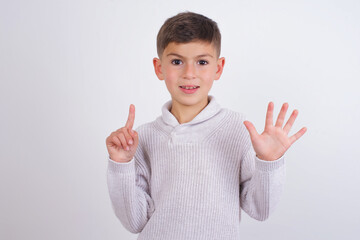 Cute Caucasian kid boy wearing knitted sweater against white wall showing and pointing up with fingers number six while smiling confident and happy.