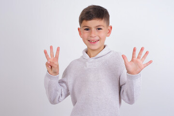 Cute Caucasian kid boy wearing knitted sweater against white wall showing and pointing up with fingers number eight while smiling confident and happy.