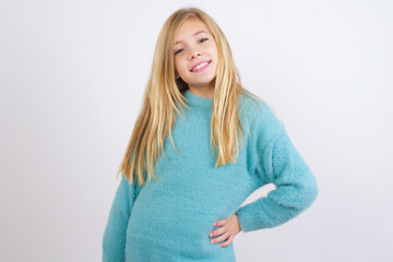 Studio shot of cheerful Cute Caucasian kid girl wearing blue knitted sweater against white wall keeps hand on hip, smiles broadly.