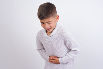 Cute Caucasian kid boy wearing knitted sweater against white wall with hand on stomach because...