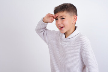 Cute Caucasian kid boy wearing knitted sweater against white wall very happy and smiling looking far away with hand over head. Searching concept.
