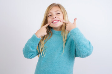 Strong healthy straight white teeth. Close up portrait of happy Cute Caucasian kid girl wearing blue knitted sweater against white wall with beaming smile pointing on perfect clear white teeth.