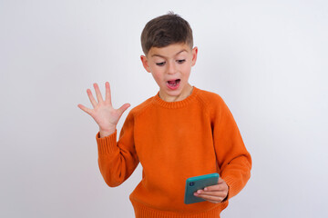 Cute Caucasian kid boy wearing knitted sweater against white wall holding in hands cell reading browsing news