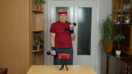 Senior elderly man watching online distance workout exercises with dumbbells on mobile phone and training, fitness, sport activity at home. Mature grandfather during coronavirus lockdown quarantine