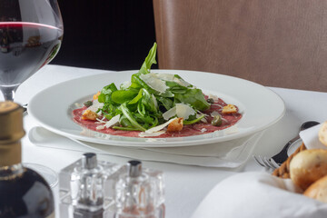 Beef carpaccio with Parmigiano-Reggiano, green leaves and red wine. Fancy food in a restaurant on white table and leather chair