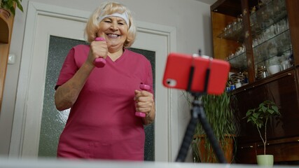 Senior elderly woman watching online distance workout exercises with dumbbells on mobile phone and training, fitness, sport activity at home. Mature grandmother during coronavirus lockdown quarantine