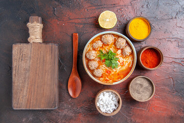 Above view of meatballs soup with noodles in a brown bowl lemon spoon different spices and cutting board on dark table