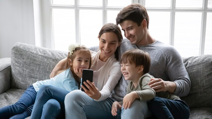 Close up happy parents with two kids using smartphone, sitting on cozy couch at home, smiling mother and father with adorable little daughter and son looking at phone screen, family making video call