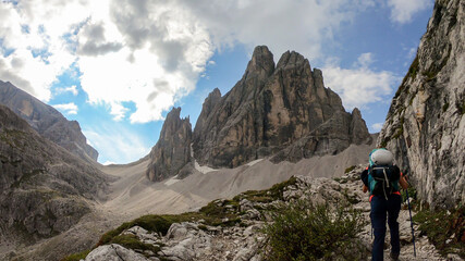 Obraz premium A woman with big backpack and a helmet hiking along a narrow pathway in Italian Dolomites. There is a massive mountain, with very steep and sharp slopes. Some smaller mountains around. Raw landscape
