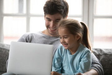 Close up happy father and little daughter using laptop together, sitting on cozy couch at home, smiling dad and adorable preschool girl looking at screen, chatting or shopping online, watching video