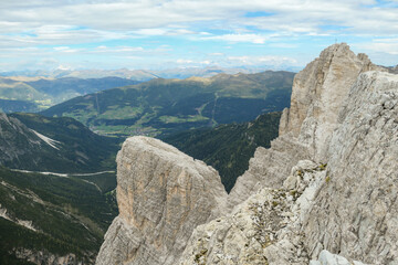 A panoramic view on a valley in Italian Dolomites. There is a massive mountain on the side, with very steep and sharp slopes. In the back there are smaller mountain chains, overgrown with green plants