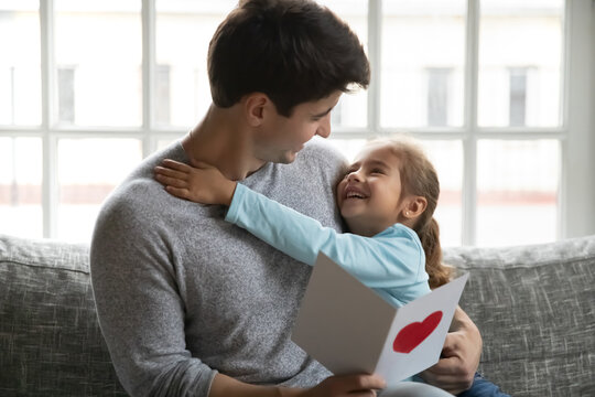 Close up smiling loving little daughter congratulating father with birthday or fathers day, happy dad hugging adorable child girl, holding greeting card with red heart, sitting on cozy couch at home - Powered by Adobe