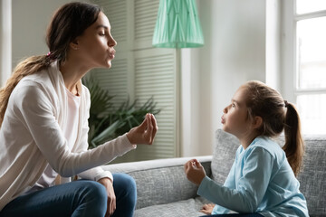 Close up little girl involved in speaking lesson with professional therapist, sitting on couch at home, caring mother teaching adorable daughter correct pronunciation, working with speech defects