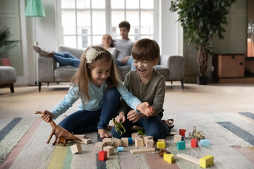 Close up happy kids cute sister and brother playing with colorful toys together while parents relaxing on couch, laughing happy little girl and boy sitting on warm floor with underfloor heating