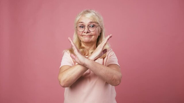 Emotional grandmother in glasses showing stop gesture, crossing hands and shaking head as rejection, pink background
