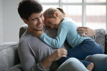 Close up happy father and little daughter cuddling and hugging, sitting on cozy couch at home,...