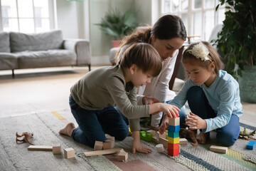 Close up mother playing with little son and daughter playing, building tower from colorful wooden blocks, sitting on warm floor with underfloor heating at home, family spending weekend together