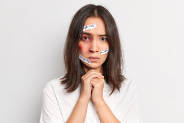 Battered bruised ethnic woman keeps hands together being afraid after abuse of violent man isolated over white background. Physically abused female with bloody eye. Stop domestic violence concept