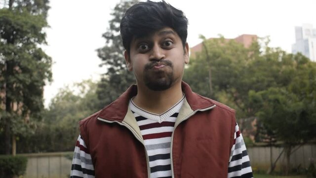 Indian boy making funny faces by puffing his mouth and cheeks with air and moving his head. He is wearing stripped Shirt   