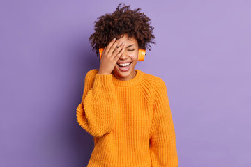 Fototapeta na wymiar Joyful positive young African American woman makes face palm wears stereo wireless headphones listens audio track poses against vivid purple background. People happy emotions and leisure concept