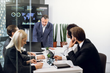Mature male chief leans on the table and talking with his team, professional successful multiethnic male and female colleagues, working together over joint strategy of company