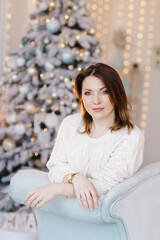 A young, attractive woman in a white sweater sits on the sofa near the Christmas tree and light