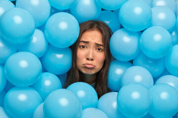 Fototapeta na wymiar Frustrated sad Asian woman purses lips looks unhappily at camera sticks head through blue balloons upset to have spoiled holiday feels lonely during her birthday. Depressed girl during festive event