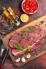 Above view of red meat on wooden cutting board and garlic green fork and knife chopped pepper fallen oil bottle on dark background