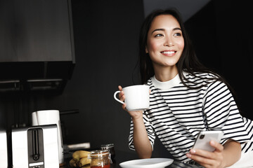 A cheerful young woman in casual clothes holds a cup of hot coffee in her hand and writes a message on the phone against the background of a home kitchen interior