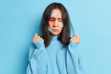 Abused assaulted woman clenches fists stands with big hematoma bruise on face expresses fear and despair being beaten closes eyes wears blue jumper poses indoor. Violence and feminism concept