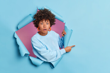 Shocked African American woman astonished by awesome advertisement indicates on right at blank space wears casual sweater poses through paper background in hole. People and promotion concept - obrazy, fototapety, plakaty