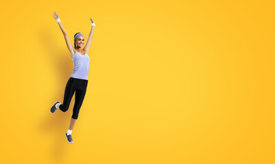 Fototapeta na wymiar Full body of joyful young woman jumping or doing fitness exercise, isolated against yellow color background. Fit girl with raised up hands, in grey sportswear, at studio shot.