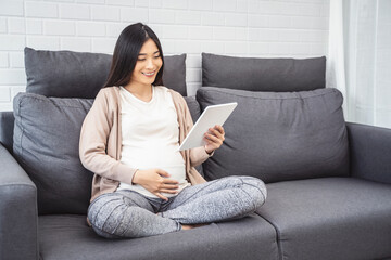 beautiful asian pregnant woman smiling using smart tablet device watching video placing hands on...