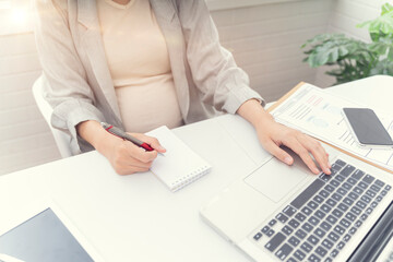 pregnant asian woman working from home in modern white office using pen writing on notepad with tablet laptop computer technology smiling with happiness of motherhood pregnancy expectant new born baby