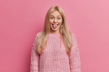 Funny blonde woman sticks out tongue and makes grimace wears warm knitted sweater poses against pink background has fun mimicks childish faces stays happy and positive. Human face expressions