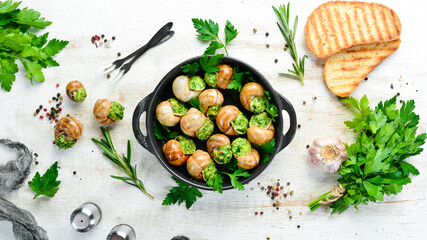 Baked snails with garlic butter and fresh herbs on a black plate on a white wooden background. Top view. Free space for your text.