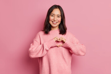 Attractive brunette young Asian woman feels happy and romantic shapes heart gesture expresses...