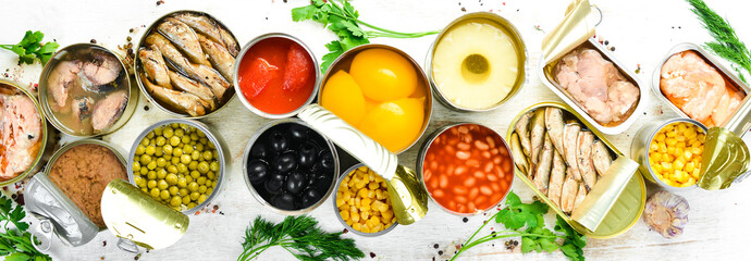 Food background in tin cans. Canned vegetables, beans, fish and fruits on a white wooden background. Top view.