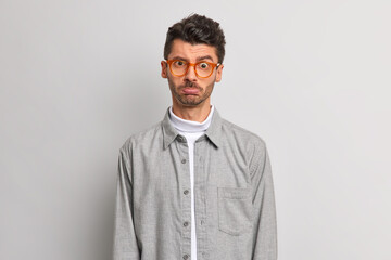 Upset offended man purses lips looks disappointed at camera expresses negative emotions stands discontent against grey background dressed in shirt wears transparent eyeglasses. Face expressions