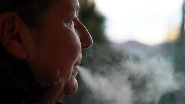 Woman smoking a cigar outdoors in the field and releasing the smoke through her nose and mouth. Woman relaxing smoking a cigarette outdoors at sunset.