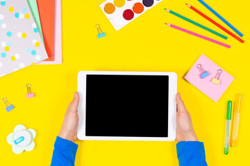 Online learning, back to school, remote education at home. Kid hands holding digital tablet computer. Headphones and school supplies on yellow background. Top view