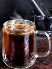 A cup of black coffee with steam on wooden table. A glass mug of hot coffee. Morning coffee. Marble mortar for grinding grains.Vertical view.