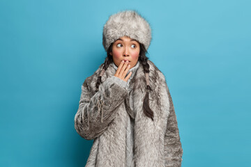 Beautiful Asian woman with two pigtails covers mouth and feels shocked wears warm natural fur coat and hat dresses for cold weather lives at north isolated over blue background. Winter female