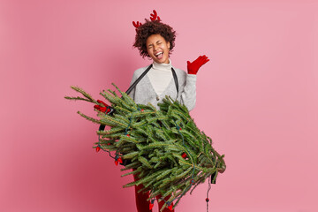 Overjoyed Afro American female model sings song along raises hand and feels amused on New Year party carries evergreen fir tree enjoys winter holidays isolated on pink background. Celebration concept