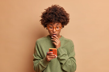 Surprised dark skinned woman stares at smartphone display checks email box reads newsfeed keeps mouth opened from wonder dressed in casual jumper isolated over brown background. Technology concept
