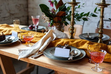 long wooden party table with stylish decoration, with candlesticks, vases of flowers, glasses with wine, plates and food in a room with a white brick wall