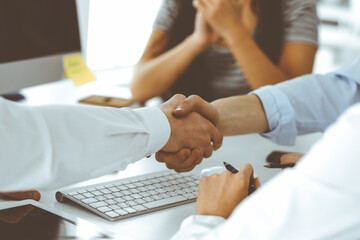 Obraz premium Two businessmen are shaking hands in office while sitting at the desk, close-up. Colleagues applauding of success meeting end. Business people concept