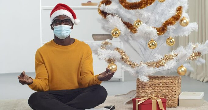 Afro american guy black man sitting on floor under christmas tree in lotus position practicing breathing exercises meditating fight stress for new year holiday hears noise opens one eye looks around